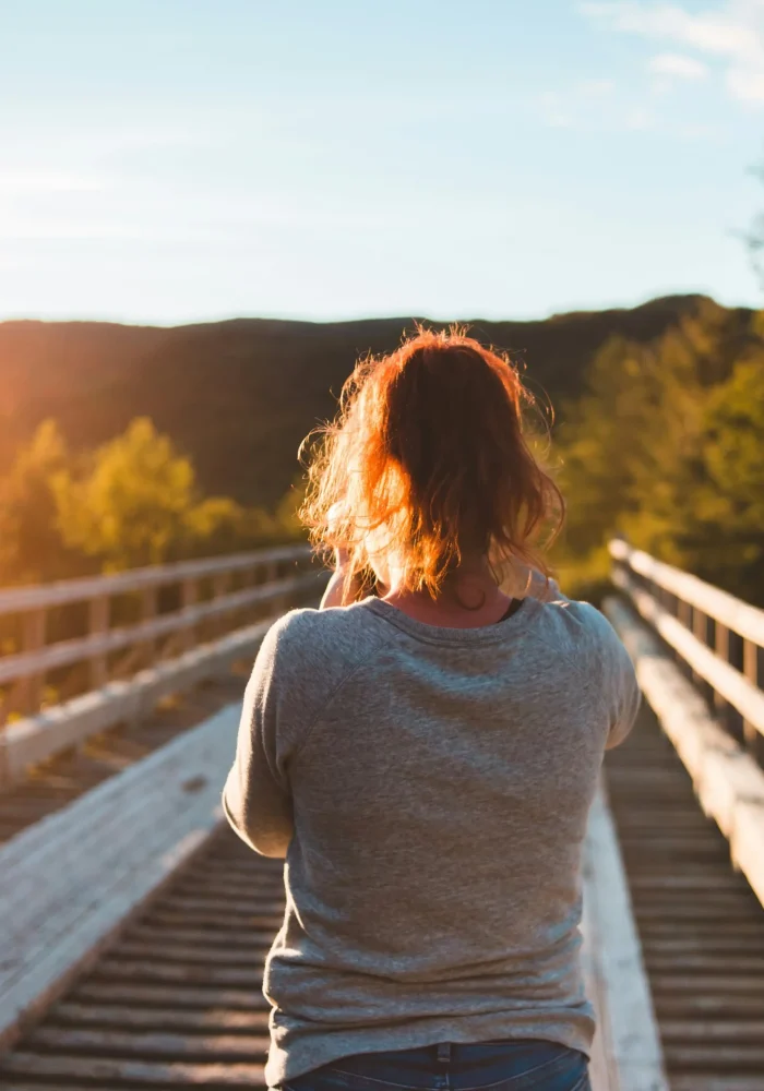 Personne de dos marchant sur un pont en bois au coucher du soleil, entourée de nature